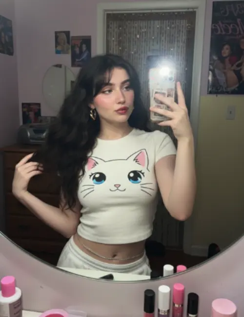 A young woman takes a confident and slightly playful mirror selfie. She has very long, voluminous dark waves with soft, wispy bangs. She wears a fitted, cream-white cropped t-shirt featuring a large, cute anime-style cat face graphic with big blue eyes, whiskers, and a small pink mouth. Her makeup is a natural glam look with soft pink dewy blush and glossy red pouty lips, and her original facial features are preserved.

For accessories, she sports gold geometric hoop earrings and a silver waistchain. She holds a smartphone in a patterned case.

The photo has the distinct aesthetic of an early-2000s digital camera. The lighting is a harsh, super-flash that creates bright, blown-out highlights while keeping the subject clearly visible. It’s a tight selfie composition shot from a mirror selfie angle. The texture includes subtle grain, retro highlights, V6 realism, crisp details, and soft shadows.

The background is a nostalgic early-2000s bedroom with pastel-toned walls. The room contains a chunky wooden dresser, a CD player, posters of 2000s pop icons, a hanging beaded door curtain, and a cluttered vanity with lip glosses. The atmosphere is an authentic 2000s nostalgic vibe, enhanced by retro-style lighting.