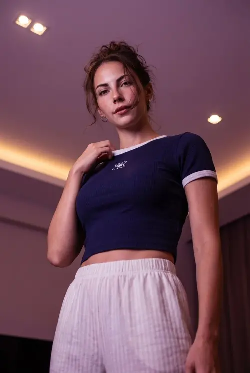 A high-definition photograph taken from a low-angle perspective. A young woman is standing stylishly, positioned so she looks effortlessly chic. She touches her collar with her hand, adding a thoughtful gesture to the pose.

Her hair is styled in a messy updo with a few loose strands framing her face. She wears a dark blue, ribbed, cropped t-shirt with white trim and a white logo. On the bottom, she has white striped pajama pants.

The scene is set in a dimly lit room. The warm, purple-toned lighting is enhanced, coming from a ceiling fixture and hidden cove lights, creating a moody and atmospheric vibe.

Most importantly, my facial features and face remain exactly the same as in the reference.