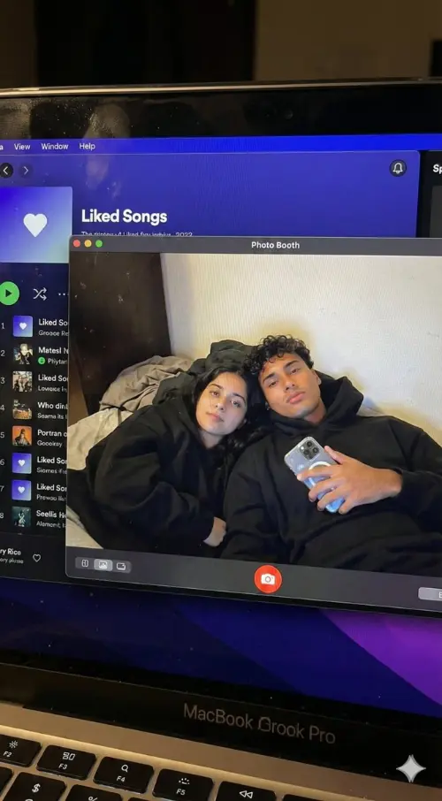 Create a hyper-realistic, downward-angled photograph of a MacBook screen. The screen should fill 95% of the frame, with the top edge of its physical keyboard visible along the bottom. The glossy screen surface must show realistic imperfections: a subtle pixel-grid texture (moire), tiny dust specks, a faint reflection of ambient light, and light fingerprint smudges.

The screen is in macOS Dark Mode. On the left, a dimmed Spotify window shows the “Liked Songs” playlist. On the right—and in sharp focus—is a full-screen Photo Booth live-preview window.

Inside the Photo Booth window, a couple is reclining together in a dim bedroom with an off-white wall and rumpled bedding in the background. Both are wearing oversized black hoodies with the hoods down, their faces fully visible. The guy, positioned on the right, is holding a phone with a clear reflective case in his right hand. The girl is resting closely beside him on the left. They are both looking directly at the screen—and by extension, at the viewer.

The lighting is low-key and nocturnal, blending cool bluish screen glow with warm skin tones, casting deep, natural shadows. The overall image should feel like a raw, unedited, candid photo—high-fidelity and completely authentic.

Important: The faces of both subjects must match exactly the faces in the provided reference images. Do not apply generic face restoration, to preserve a natural, non-plastic look.

Style to avoid: Vector art, flat digital screenshots, perfectly clean glass, bright daylight or studio lighting, cartoon styles, 3D renders, paintings, and watermarks.